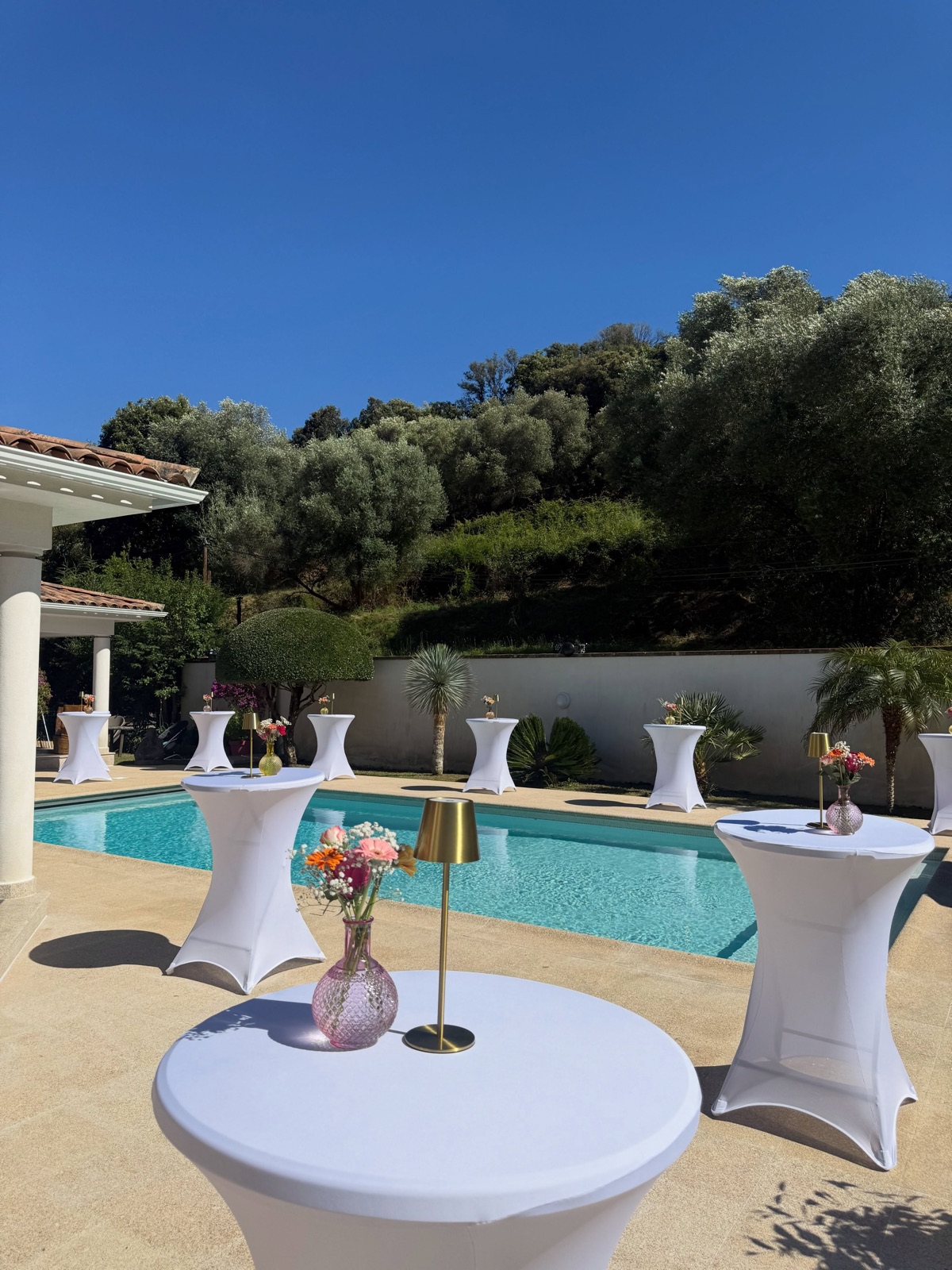 Réception en plein air au bord d’une piscine en Corse, tables mange-debout habillées de blanc